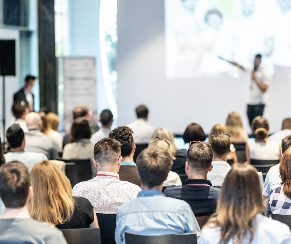 A room full of people listening to a speaker on stage at a conference.